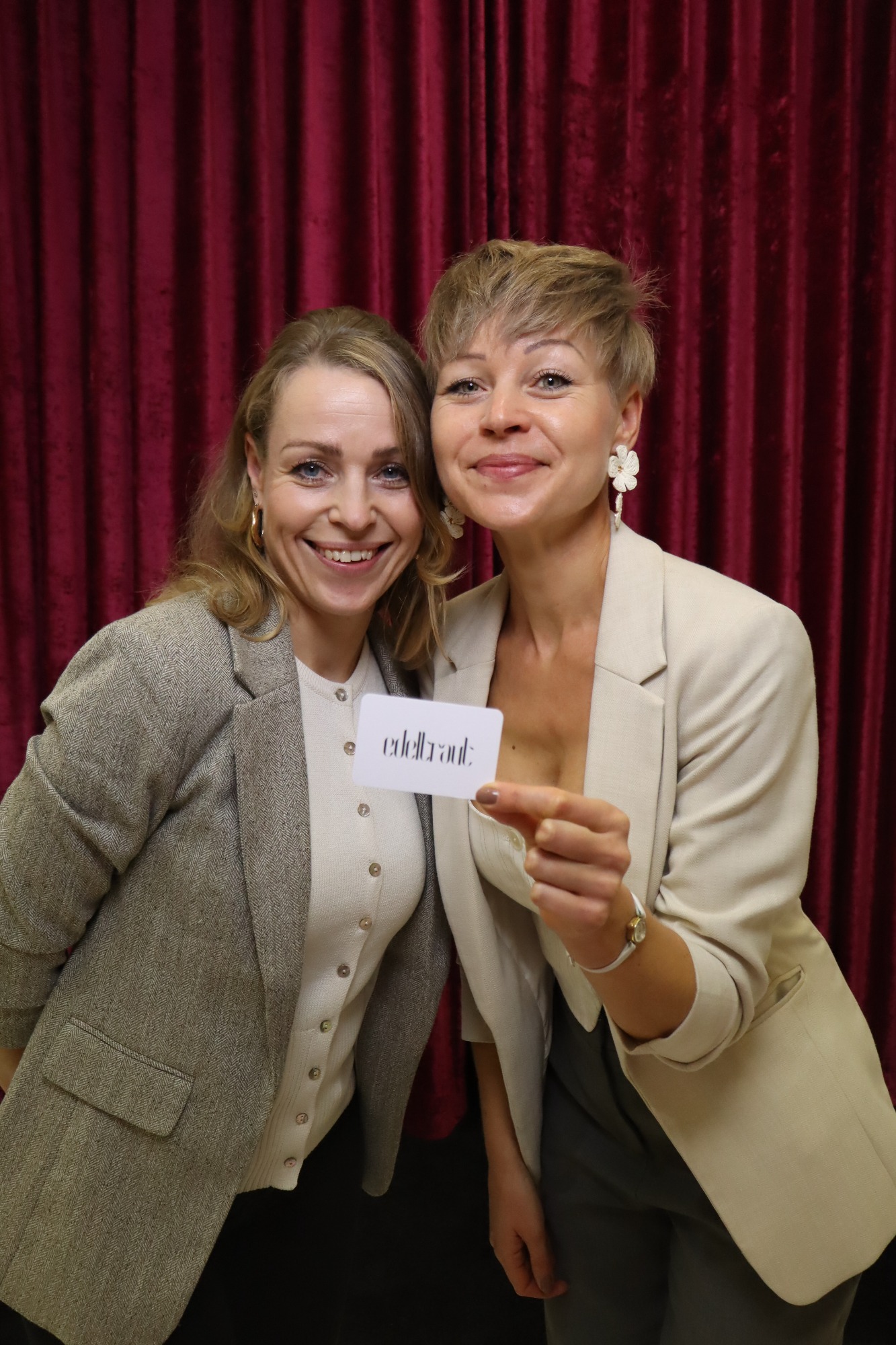 Zwei Frauen in Blazer lächeln vor einem roten Vorhang, eine hält eine Karte hoch.