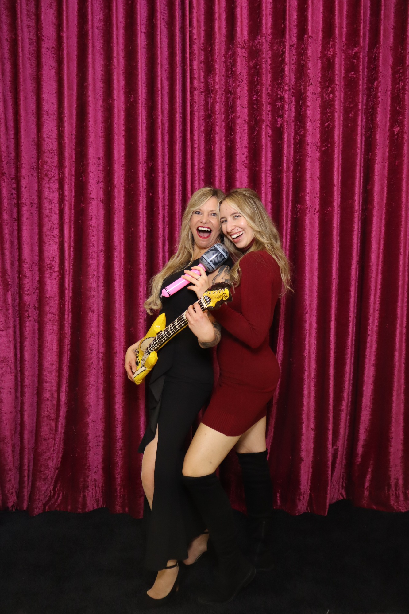 Zwei Frauen posieren fröhlich vor einem pinken Vorhang, eine hält eine Gelbgitarre.