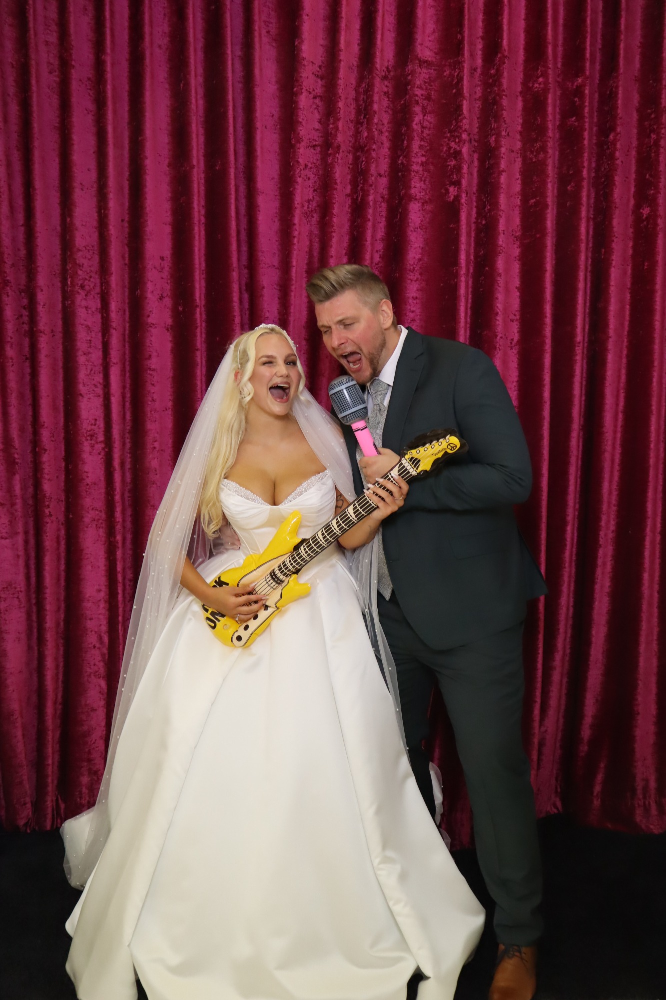Braut mit Gitarre, Bräutigam daneben, lächeln vor pinkem Hintergrund.
